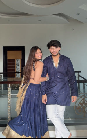 A couple posing indoors in traditional South Asian attire. The woman wears a navy blue sleeveless lehenga choli with gold border detailing, and the man wears a matching navy kurta with white trousers.